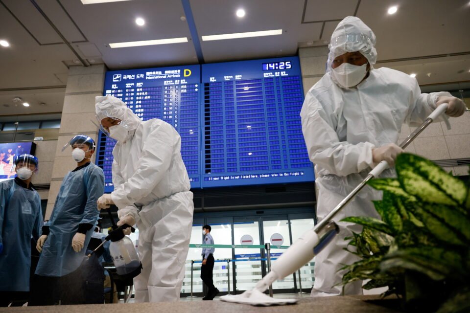 Workers Wearing Protective Gear Disinfect An Arrival Gate As An Electronic Board Shows Arrivals' Information At The Incheon International Airport, In Incheon